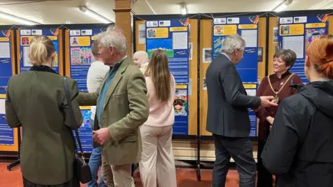 ANDREA PLUCK/BBC Several people gathered in a library hall wearing formal clothing. Around them are displays of drawings and letters written by children