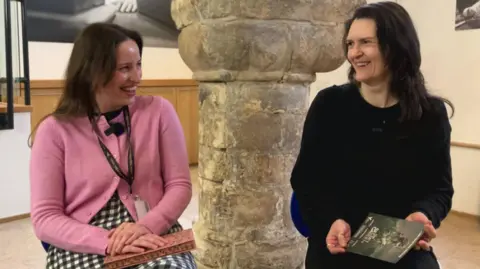 BBC/Seb Cheer Catrin and Elizabeth look at each other, laughing. They are sitting on chairs in front of the camera.