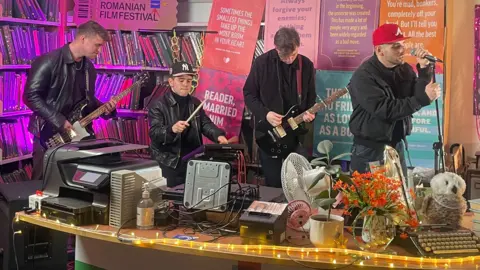 Bass, drums, guitar and vocals for a four piece band standing behind a library information counter