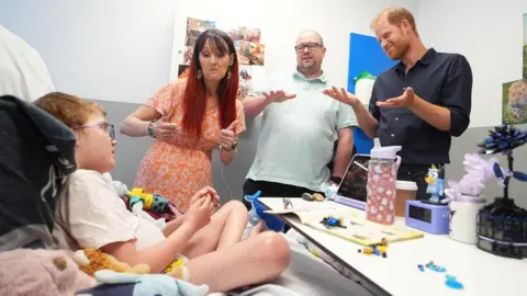 PA Media Prince Harry meets a young child in hospital. He stands by the bed with the boys parents. 