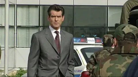 Pierce Brosnan, who played James Bond, stands outside an office building. He is wearing a grey suit and has thick, dark brown hair. He is frowning slightly and has a hand in his pocket. There are people in camouflage uniforms facing away from the camera and an emergency car which says 'Polis' on it.