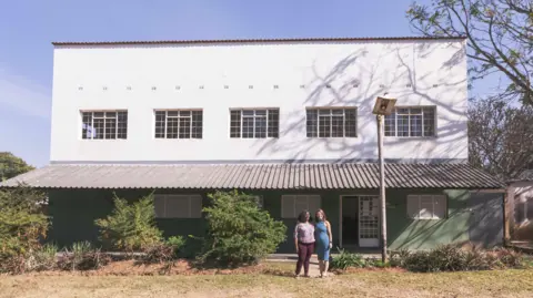 GoGo Olive Two women stand outside a two-storey white building. There are five windows on the first storey, while a tin roof provides shade for a paved area outside the ground floor. 