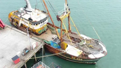 The Cornishman trawler pictured moored to a dock. It has a white cabin with a large deck. The vessel is suffering from rust in most areas. 