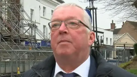 BBC A man with white hair and glasses in a dark coat, white shirt and blue tie stood in front of a building with scaffolding