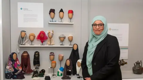 St Albans Museums A woman with a green headscarf standing in front of a museum exhibit featuring a collection of traditional headwear displayed on mannequin heads. The headwear includes various styles, fabrics and colours, such as scarves, turbans and caps.