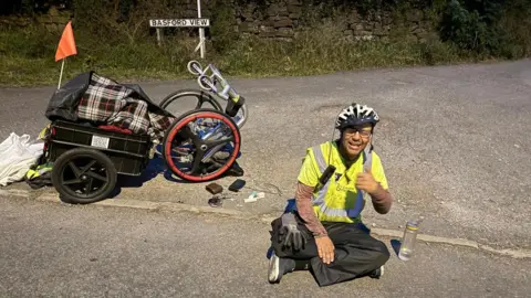 A man wearing a yellow high visibility jacket is sat on a pavement. Behind him is a wheelchair and a trailer. The wheelchair has been tipped over. A road sign behind the wheelchair identifies the location as Basford View.