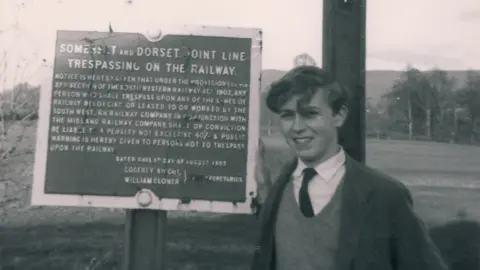 Black and white photo of Paul Antell as a teenager standing next to a sign which explains it is forbidden to trespass on the Somerset and Dorset line.
