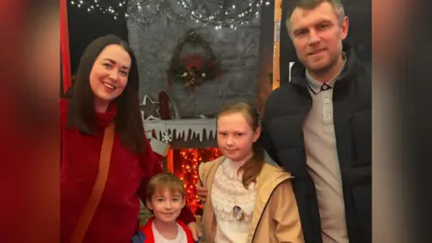 Rachel Taylor Rachel is on the left of the image wearing a red jumper, she has long brown hair. Beside her is a small boy with light brown hair wearing a white T-shirt and beside him is an older girl with a brown ponytail wearing a khaki raincoat over a cream fairisle sweater. On the right is a man with grey/brown hair wearing a dark jacket over a grey polo shirt.