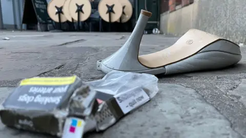 BBC/Jo Makel A discarded high-heeled shoe lies on a pavement next to an empty cigarette pack and small debris. In the background, outdoor tables are folded up outside a venue.