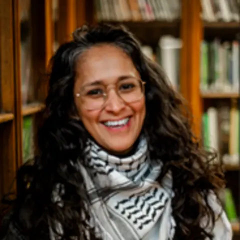 Women of the World/Joanne Coates Rumana Yasmin has long black wavy hair and glasses. She is smiling and wearing a black and white scarf. There are shelves behind her with books.