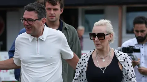 EPA Andy Burnham wears a white polo top with a zip and black glasses. He is walking with his wife Marie-France van Heel, and has a hand on her back.
Van Heel has light blonde hair and wears large black sunglasses, thick silver jewellery and a black vest with an open white shirt which has black polka dots on it.