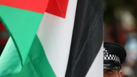 A green, red, white and black Palestinian flag drapes down, obscuring the face of a police officer. The man wears his black police hat, which has a black and white checkered detail around it.