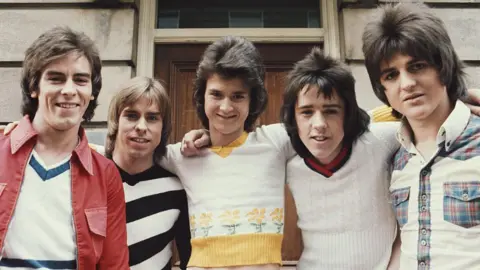 The five members of the Bay City Rollers pose arm-in-arm for the camera. Alan wears a trendy mullet with a white sweater and a red jacket with large lapels, Derek, also sporting a mullet, wears a black and white striped jumper, Les, in an impressive bouffant hairstyle, wears a tight white sweater with yellow flower trim, Woody leans forward with his spiky mullet and a white cricket jumper, and Eric keeps it tartan with a white, tartan-trimmed shirt and an impressively layered face-framing hairstyle. They stand outside with a closed wooden door behind them.