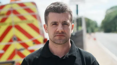 John Fairhall/BBC Sam Dimond has brown hair and is wearing a black polo shirt. Behind him is a yellow and red traffic van.