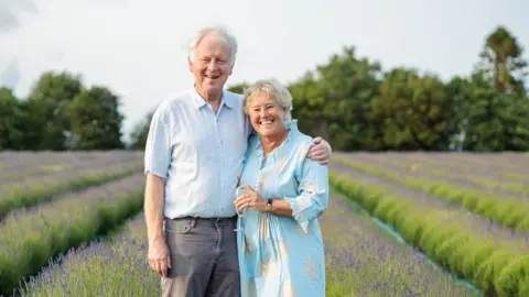 Ian Jamie owns the farm with his wife Pippa and the couple are seen here celebrating their 40th wedding anniversary with a glass of bubbly in a lavender field. Both are wearing light blue and they are standing together looking happy.