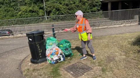 John Devine/BBC Margaret Faulkner carrying a litter grabber. A rubbish bin is beside her. Also in the background are some metal railings beside a roadway. There is a pile of full green bin bags beside the public bin.