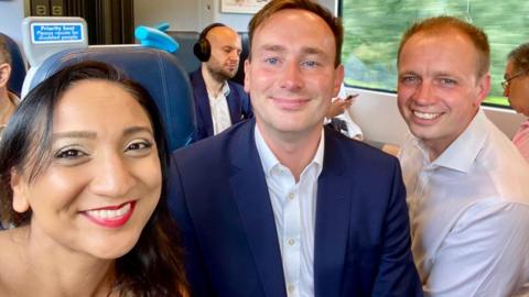 Tom Hayes in the centre with a navy suit and white shirt - no tie. He has short brown hair. To the right is Darren Paffey in a white, long-sleeved shirt with no tie. He has shirt, fair hair and is clean shaven. Satvir Kaur is to the left with long dark hair. All are smiling at the camera