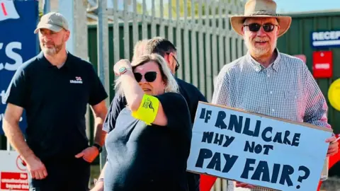 Three protesters stand outside a large metal gate. In the centre, a woman with blonde hair and sunglasses is wearing a bright yellow "Unite" armband. To her left, a bearded man is wearing a black polo shirt with "Lifeboats" printed on one side of the chest and a green baseball cap. To the right, a man in a checkered shirt donning sunglasses and a cowboy hat is holding a sign to the camera; it reads: "If RNLI care why not pay fair?"