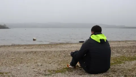 A young man sat on the side of a water body looking out at the gentle waves lapping on the shore.