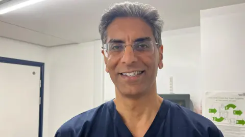 Dr Saeed Ahmed commonly known as Sid smiles at the camera at the Renal Department at Sunderland Royal Hospital. He is wearing blue scrubs and is wearing glasses with a silver frame. 