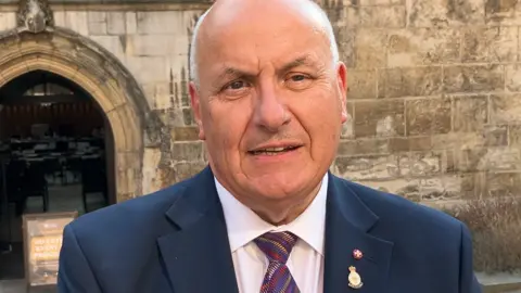 BBC/Victoria Scheer Close-up of a man standing outdoors in front of a historic stone building. He is facing the camera and wearing a dark navy suit jacket, a white shirt and a striped tie. Two small lapel badges are also visible on the jacket.