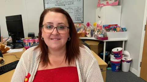 A woman wearing red and white and glasses is looking at the camera. She has long dark hair and is prominent in the image, which also includes a whiteboard and collecting buckets on the floor in the background.