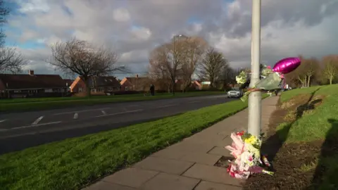 View of the scene of the crash. Some flowers and balloons are tied to a lamppost next to a road, with grass verges either side and houses beyond.