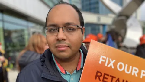 A man wearing glasses holds an orange sign that is mostly out of shot, the words "fix our" are visible. The background is out of focus but it is clear that he is standing with a group of people in front of a set of buildings