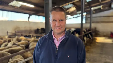 Farmer John Martin is standing in a large shed with sheep in one pen and cows in another pen. John is wearing a navy hoodie with a pink and blue stripped shirt. He has short. brown hair.