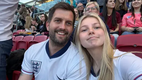 Richard Richard and Imogen wearing England football tops sitting in the stands at a football stadium taking a selfie. They are both smiling at the camera. She has long blonde hair which is straight while he has dark hair and a short beard.