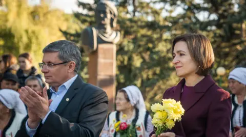     Matthew Goddard President Maia Sandu holding small flowers while visiting the campaign. He is wearing a purple suit jacket. The man standing on the left exclaims. There are people in traditional clothes in the background.