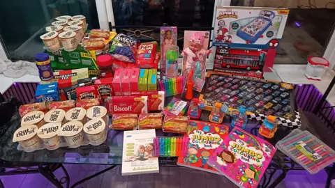 Family handout A collection of children's toys including sets of cars, colouring books, felt tip pens and food and snacks such as soups and crisps on a table in a home.