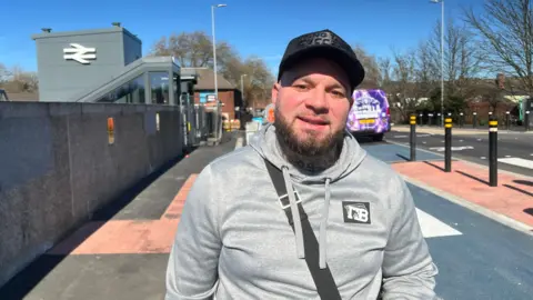 A man wearing a grey hoodie and a black cap stands outside a railway station entrance. The National Rail double arrow logo is visible on the station building behind him. A purple bus and residential street can be seen in the background on a sunny day.
