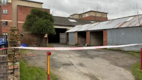 Some outdoor buildings stand on the right hand side of the photo with corrugated sides. They are attached to another building with a tree in front of them. Police tape cane be seen at the driveway entrance to all the properties