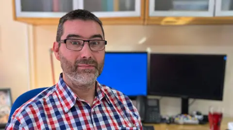 BBC Dr Dan Lane sits at a desk in his consulting room. He has short black hair and a neatly trimmed beard and is wearing a red-and-blue checked shirt. Behind him are two computer monitors.