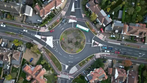 Aerial view of a roundabout. A red cycle lane is visible on roads to the left and right and on the roundabout itself. 