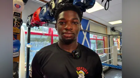 Zac Sherratt/BBC Judah Armah at the Odyssey Boxing Club in Stanwell, Surrey. Judah is wearing a black t-shirt with the club logo, and has black hair. He is standing in a boxing ring. 