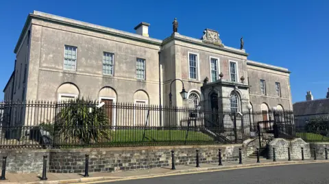 Downpatrick Crown Court - pictured on a sunny day - the court house is grey stone and surrounded by a black fence and small lawn. 