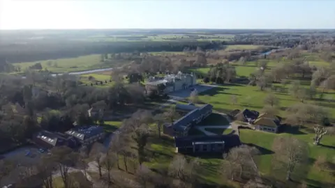 A large grey brick 18th-century stately home surrounded by trees, fields, and smaller, newer buildings.