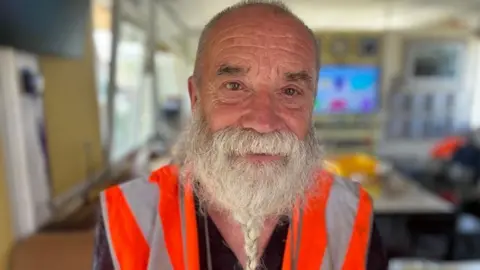 Head and shoulders of Brian Haworth inside the box which is blurred in the background. He has very short hair and a long beard which is plaited. He is wearing an orange high vis vest
