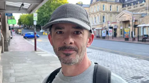 A man with a grey beard and grey jumper wearing a grey hat and black backpack. He is standing in front of a quiet road.