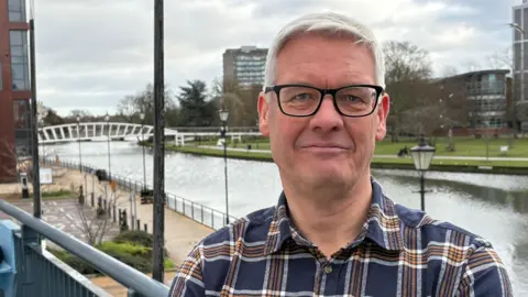 Ben Schofield/BBC Simon White standing outside and looking directly down the camera. He has grey hair, parted on the right hand side as we look at him. He is wearing black rimmed, rectangular glasses and a casual checked shirt. He is smiling slightly. Behind him is a river flowing through a low density urban environment. On the left, in the middle distance, a white footbridge can be seen crossing the river. The near bank has a light-coloured, paved footpath, while the far bank appears to be grass covered. There are a couple of low rise buildings on both the far and near banks. 