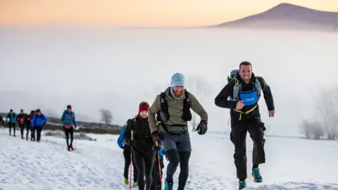 Racers run and trek through snow holding walking poles. They wear warm clothing. A mountain can be seen in background.