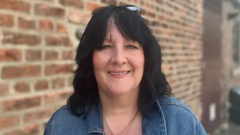 Steve Jones/BBC Jenny Bryant, a 50-year-old woman who has black hair and blue eyes. She is wearing a denim jacket and smiling in the headshot photo.