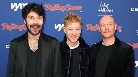 Getty Images Simon Neil, James Johnston and Ben Johnston, of the Scottish rock band Biffy Clyro, stand against a blue backdrop. They are all dressed in black t-shirts and jackets and are smiling at the camera.