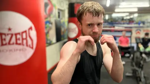 BBC A young white man in a black, sleeveless top strikes a boxing pose, with hands up, in front of a red punching bag. He is pictured in a gym.