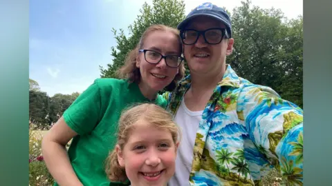 Alex Cummings A family of a man, woman and their child. The man is wearing a hawaiian shirt and a blue cap, and is smiling at the camera
