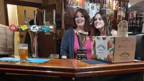 Perry St Social Club Two women behind a bar with pirate decorations, beer pumps and a pint on the bar, they are smiling at the camera