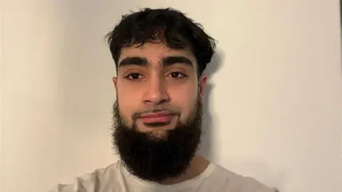 Umar Fayyaz Umar Fayyaz, a man with a thick black beard and black hair is standing against a cream-coloured wall.