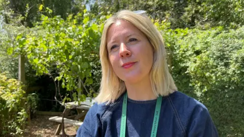 Louise Moreton, the horticulturalist for the school, is smiling and looking off to the left of the picture. She has blonde hair which is shoulder length and ihas sunglasses on top of her head. She is wearing a blue denim dress. Behind her, are a number of green plants and trees and a wooden bench. 